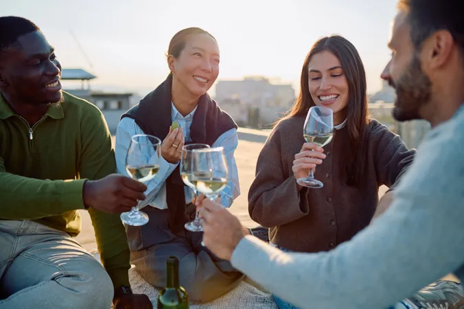 Vier Menschen sitzen im Freien, lächeln und stoßen unter klarem Himmel mit Gläsern Weißwein an, im Hintergrund ist eine verschwommene Stadtkulisse zu sehen.