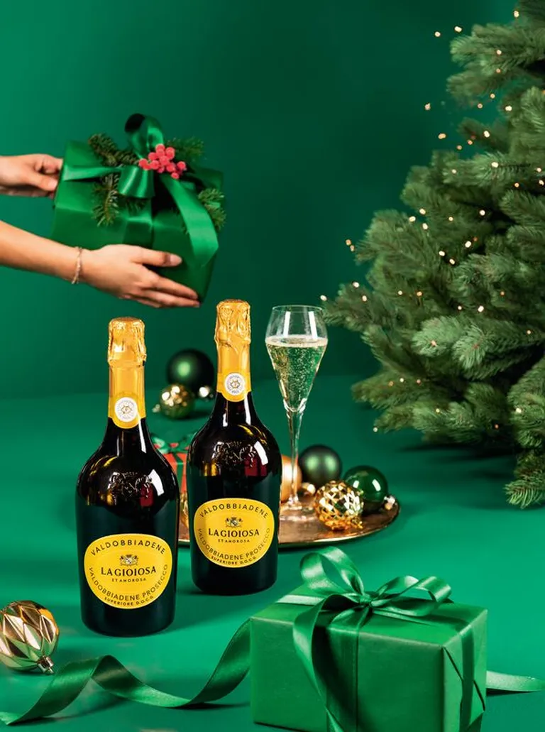 Eine Person nimmt ein grün verpacktes Geschenk, daneben stehen 2 Flaschen La Gioiosa und ein glas Sekt sowie ein Christbaum