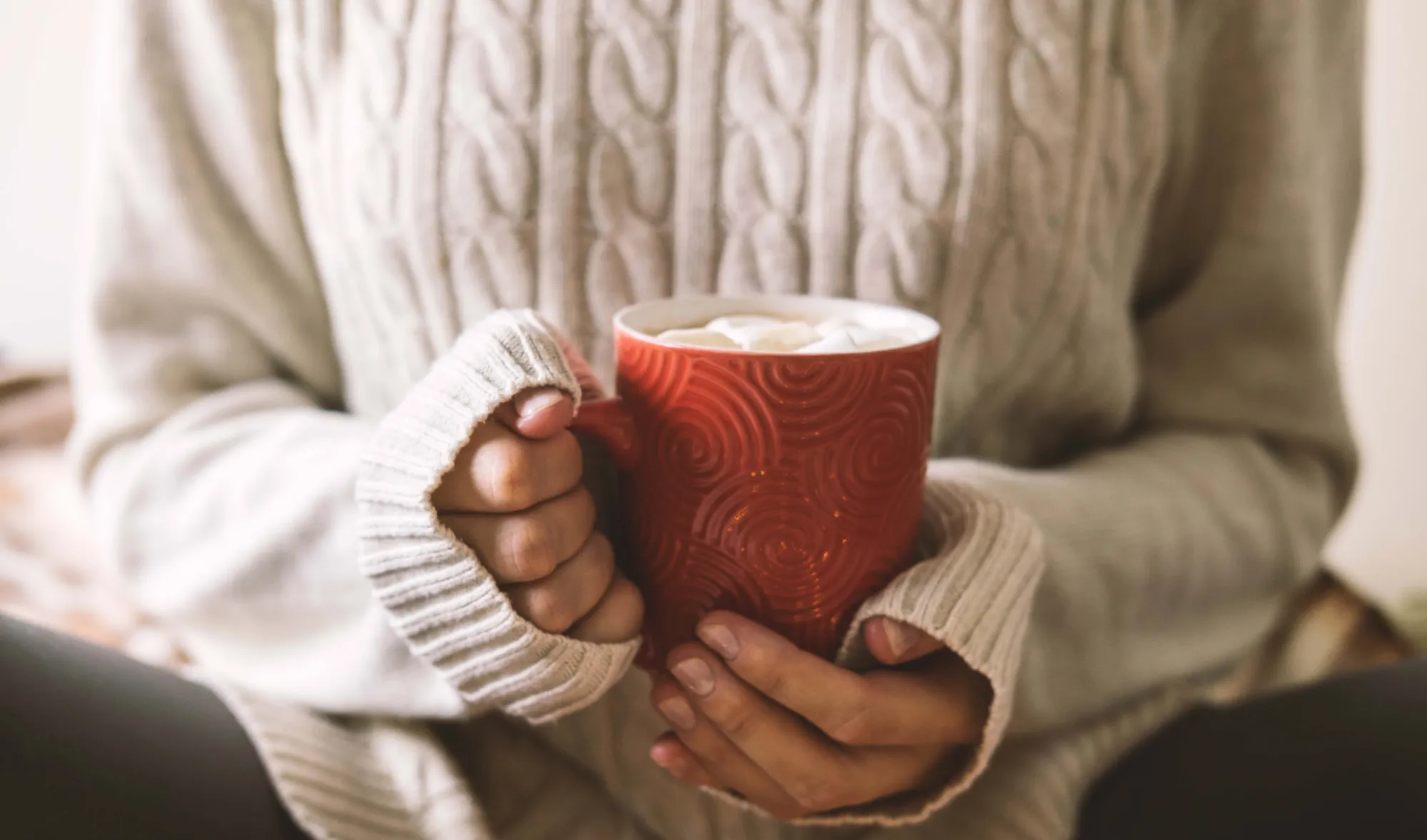 Eine Person in einem hellgrauen Pullover hält eine rote Tasse in den Händen