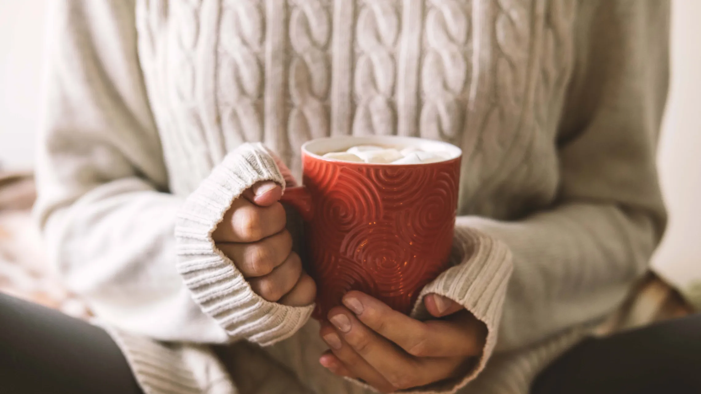 Eine Person in einem hellgrauen Pullover hält eine rote Tasse in den Händen