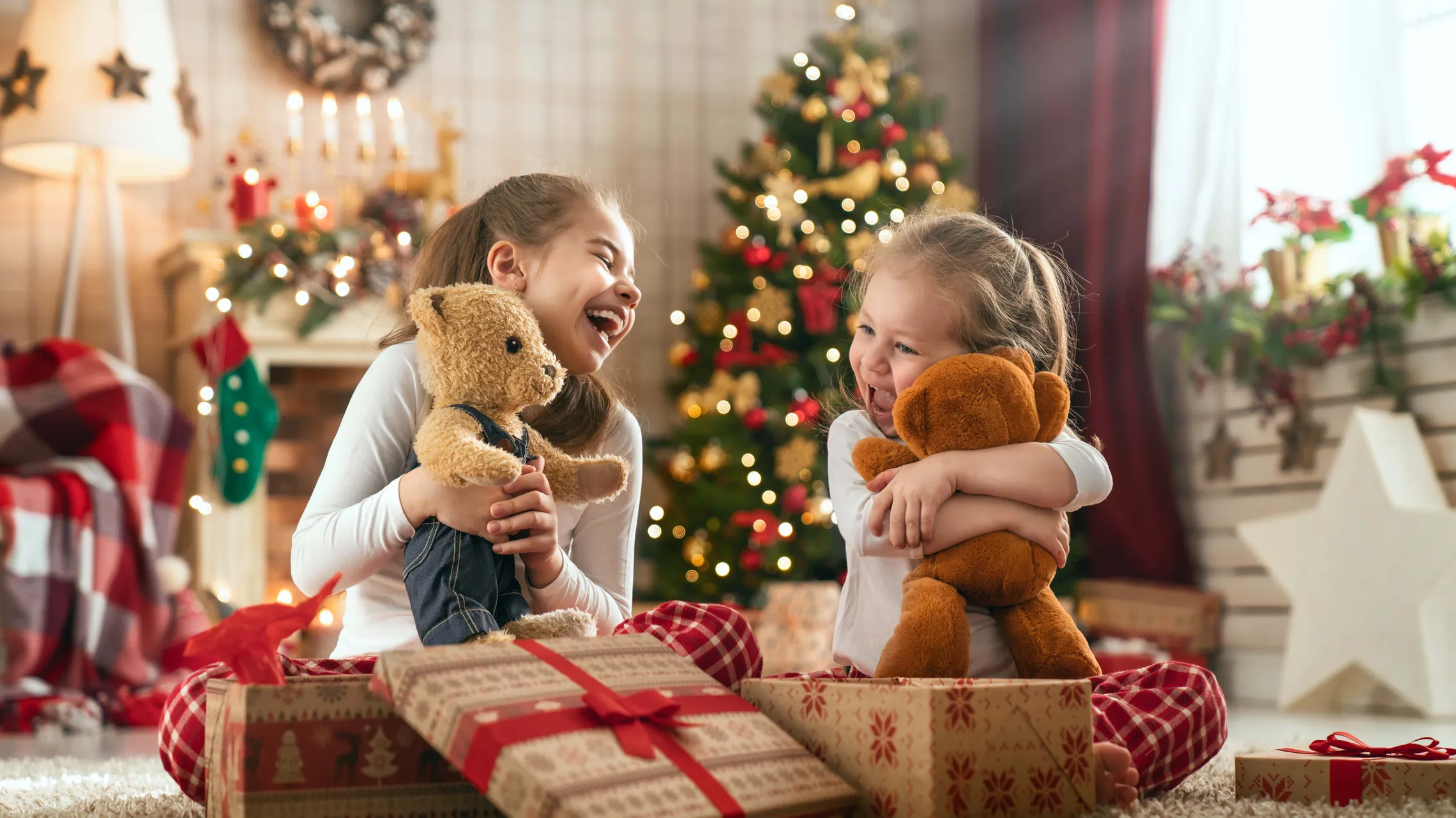 Zwei lachende Mädchen sitzen vor einem geschmückten Christbaum und freuen sich über ihre neuen Teddybären. Vor ihnen liegen weihnachtlich verpackte Geschenke