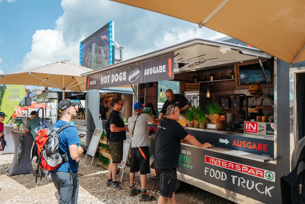 Menschen, die an einem sonnigen Tag an einem Imbisswagen bestellen, der unter einem Sonnenschirm Hot Dogs anbietet, während ein Mitarbeiter das Essen zubereitet.