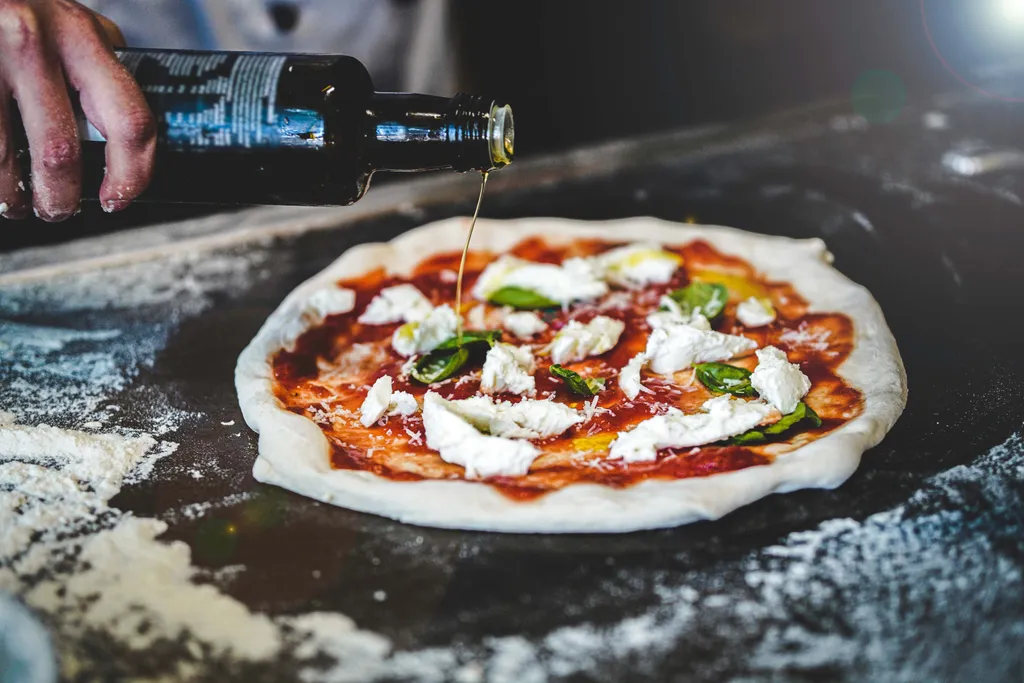 Olivenöl wird über frisch belegten Pizzateig mit Mozzarella und Tomatensauce gegossen