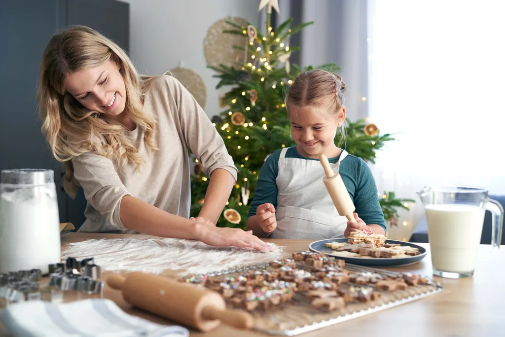 Eine Mutter und ihre Tochter backen gemeinsam Weihnachtskekse in der Küche. Im Hintergrund steht ein geschmückter Weihnachtsbaum.