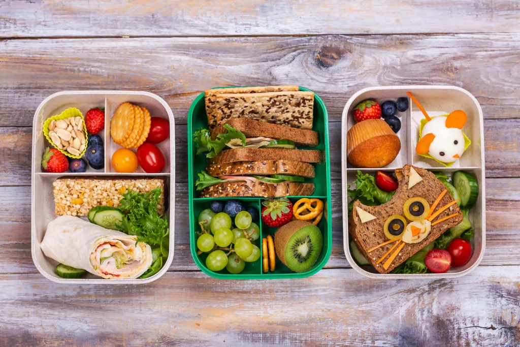 Drei Bento-Boxen mit Sandwiches, Obst, Gemüse, einem Wrap, Crackern und einem Muffin. Kreative Motive mit einer Maus und einem Katzengesicht.