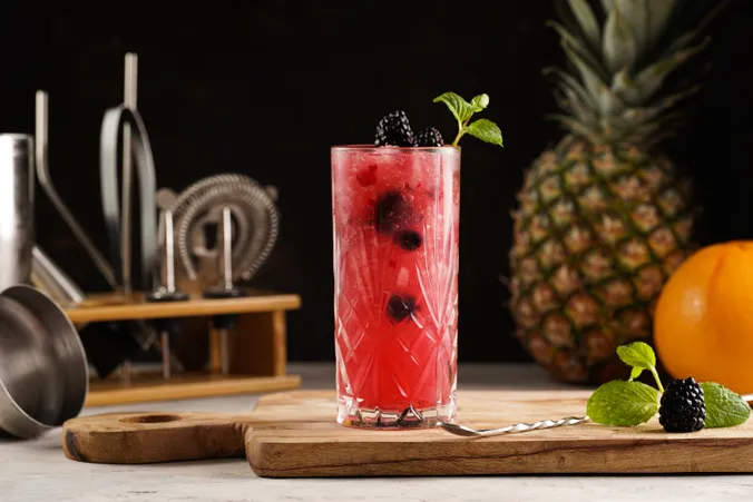 Auf einem Holzbrett steht ein farbenfroher Brombeercocktail, garniert mit Minze, im Hintergrund sind eine Ananas und Zitrusfrüchte zu sehen.