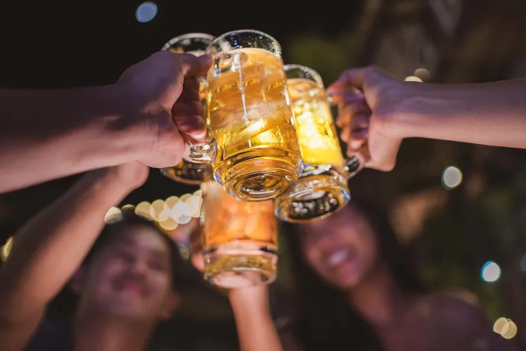 Menschen stoßen mit Bierkrügen an und feiern in der Nacht vor dem Hintergrund verschwommener Lichter.