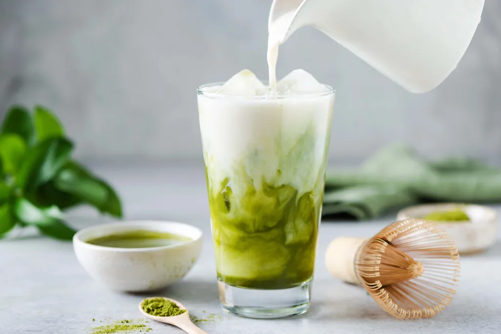 Ein gekühlter Matcha Latte in einem Glas, in das gerade Milch eingegossen wird, umgeben von Matcha-Pulver, einem Bambusbesen und einer kleinen Schale.