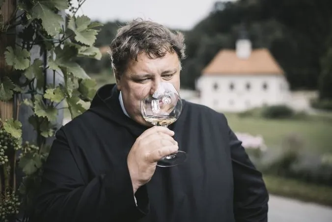 Ein Mann in einer schwarzen Robe, der an einem Glas Wein riecht; er steht im Freien inmitten von Grün, im Hintergrund ist verschwommen ein Gebäude zu sehen.