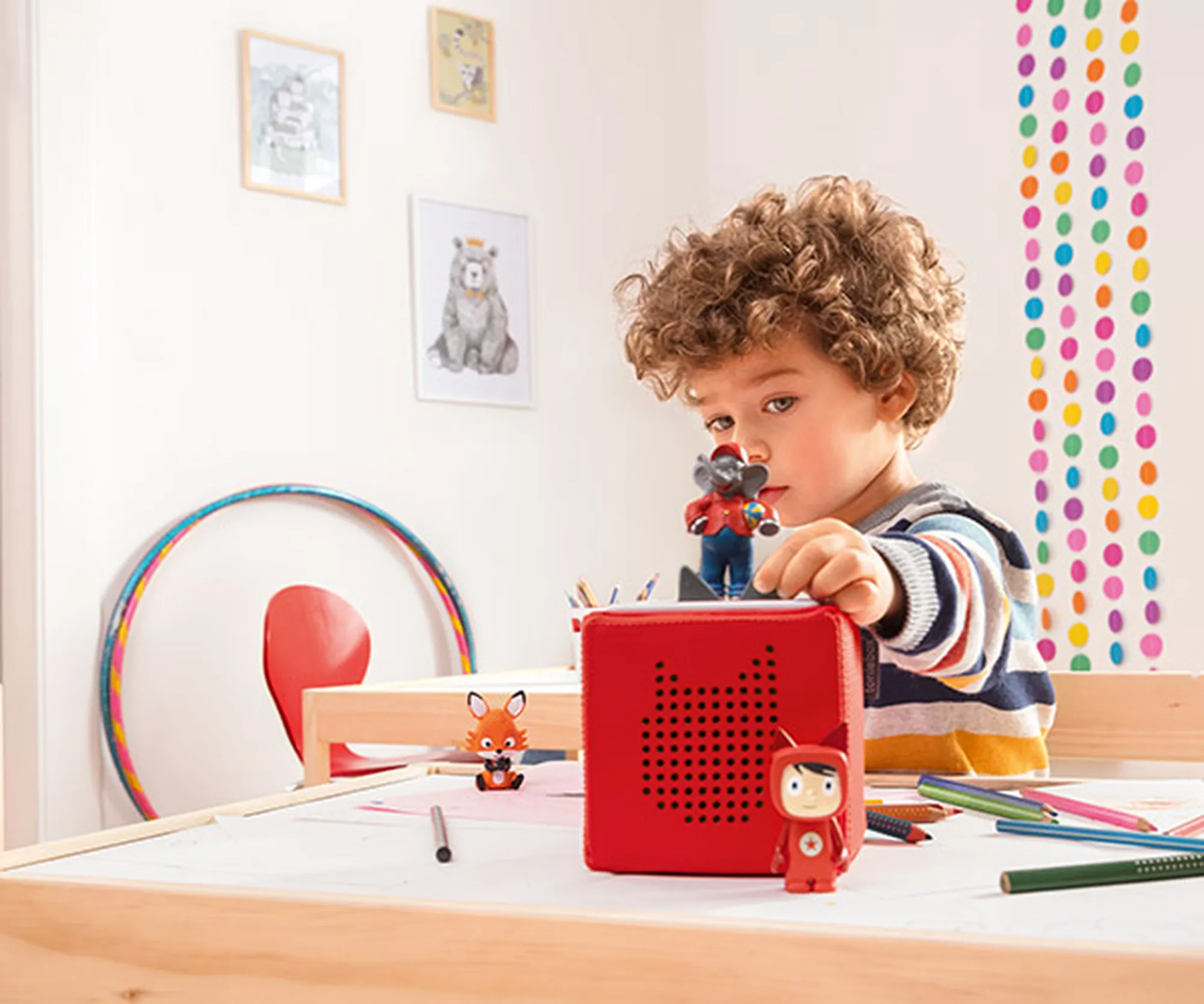 Ein Kind mit lockigem Haar spielt an einem Tisch mit einer roten Toniebox, eine Art Lautsprecher auf der man Spielfiguren platzieren kann um Musik oder Geschichten hören zu können, umgeben von bunten Zeichnungen und Mustern an der Wand.