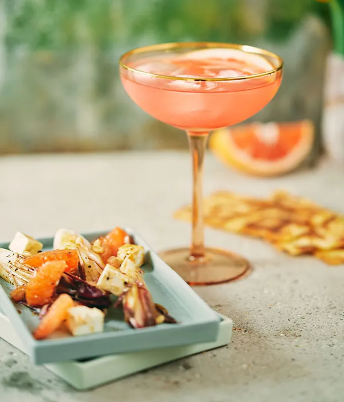 Ein rosa Cocktail in einem Coupé-Glas neben einem Teller mit gemischtem Salat, Orangenscheiben und Käse auf einer strukturierten Oberfläche.