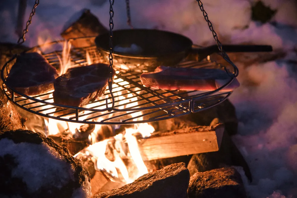Steaks auf einem Schwenkgrill über Lagerfeuer in verschneiter Winterlandschaft