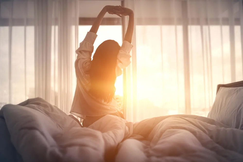 Frau sitzt morgens im Bett und streckt sich im warmen Sonnenlicht
