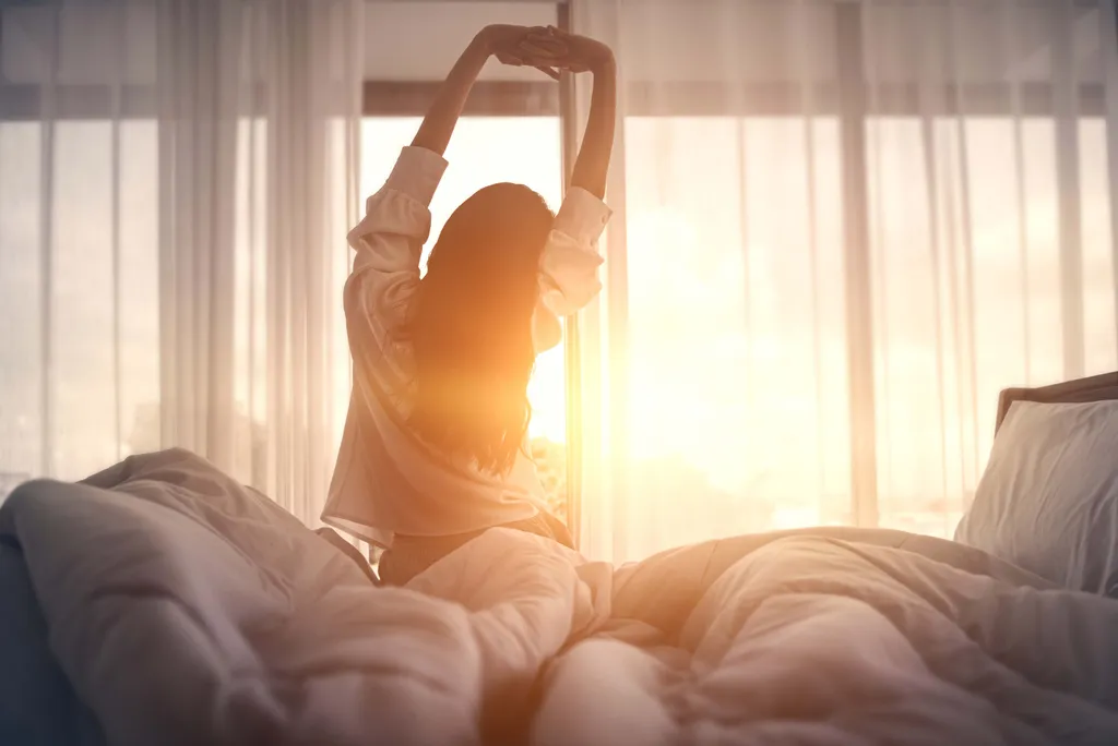 Frau sitzt morgens im Bett und streckt sich im warmen Sonnenlicht