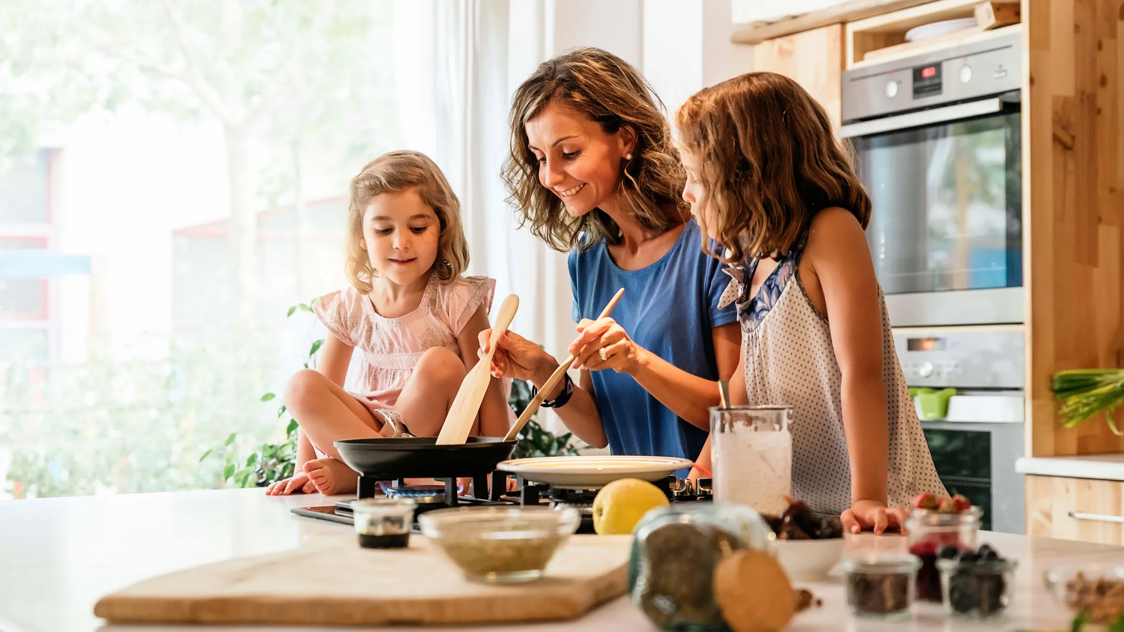 Eine Frau und zwei kleine Mädchen kochen fröhlich gemeinsam in einer hellen Küche, umgeben von Zutaten und Küchenutensilien.