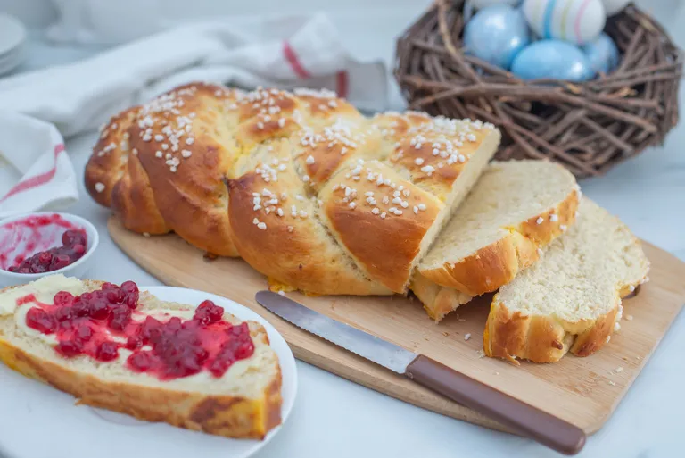 Ein Striezel mit Hagelzucker neben einem Brotmesser auf einem Holzbrett und eine geschnittene Scheibe mit Marmelade