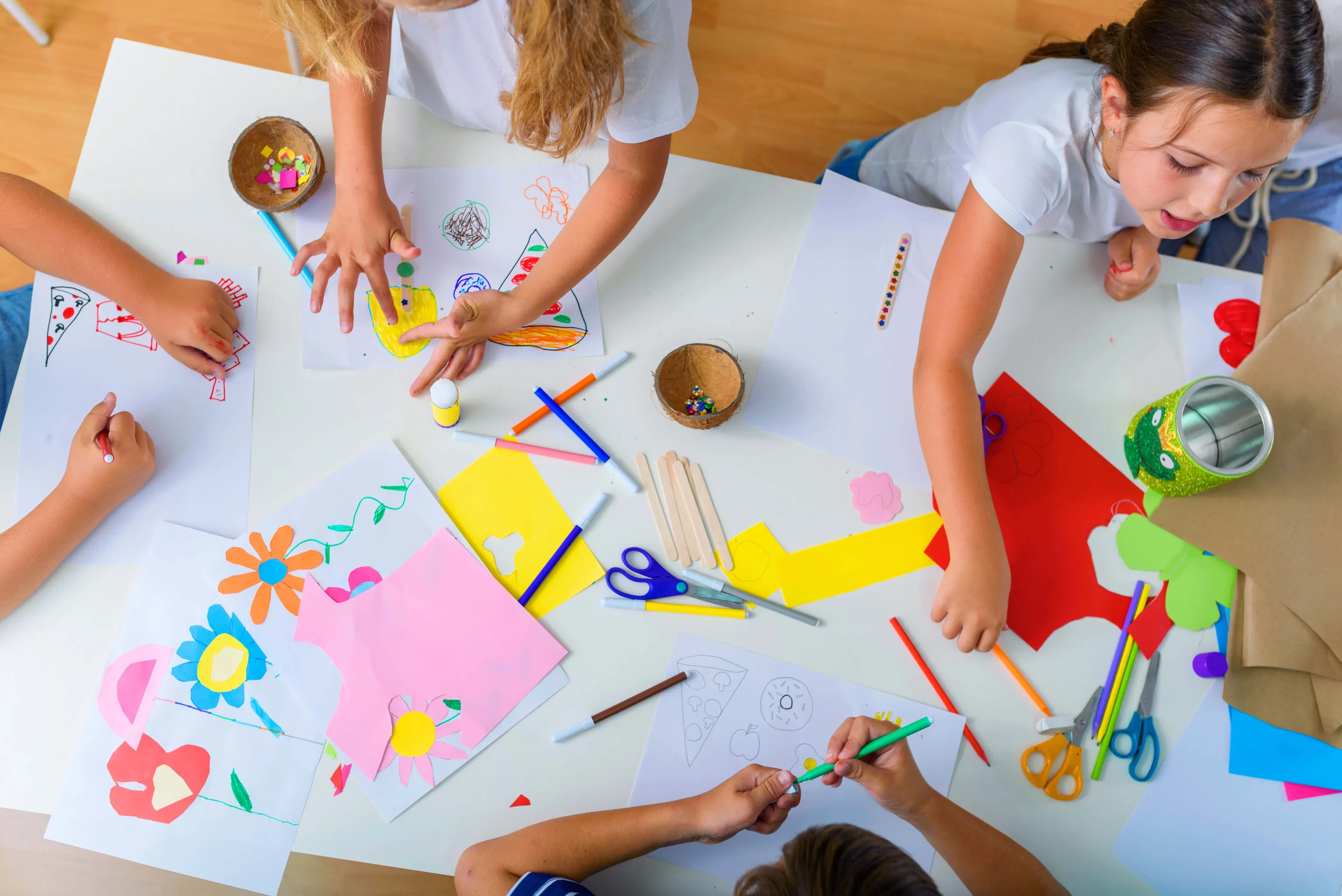 Kinder basteln an einem Tisch, malen und schneiden buntes Papier mit Schere und Klebestift aus.