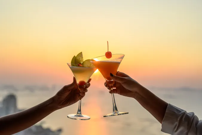Zwei Hände, die vor dem Hintergrund eines ruhigen Sonnenuntergangs über einem Gewässer mit Cocktails mit Fruchtgarnituren anstoßen.