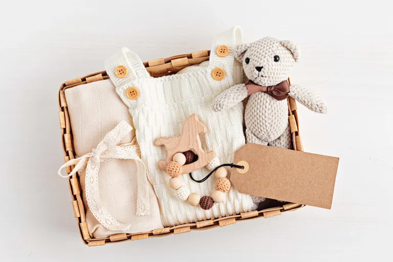 Ein geflochtener Korb mit einem gestrickten Teddybären, einem weißen Baby-Outfit, einem Holzpferd und einem beigen Geschenkanhänger vor hellem Hintergrund.