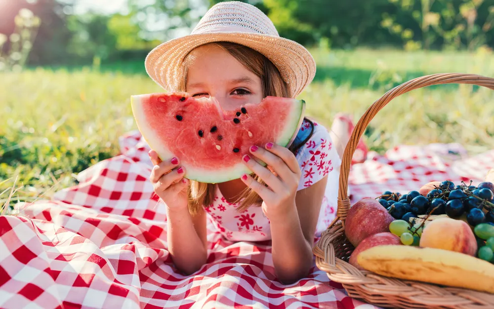 Ein Mädchen mit Strohhut liegt auf einer Picknickdecke und hält ein Stück Wassermelone ind die Kamera
