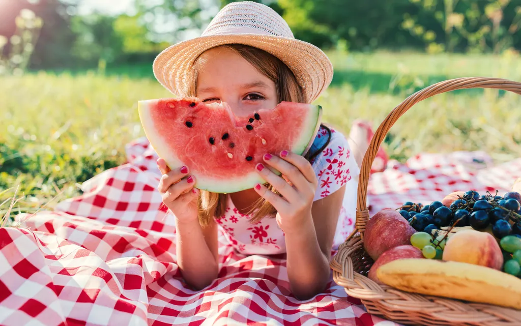 Ein Mädchen mit Strohhut liegt auf einer Picknickdecke und hält ein Stück Wassermelone ind die Kamera