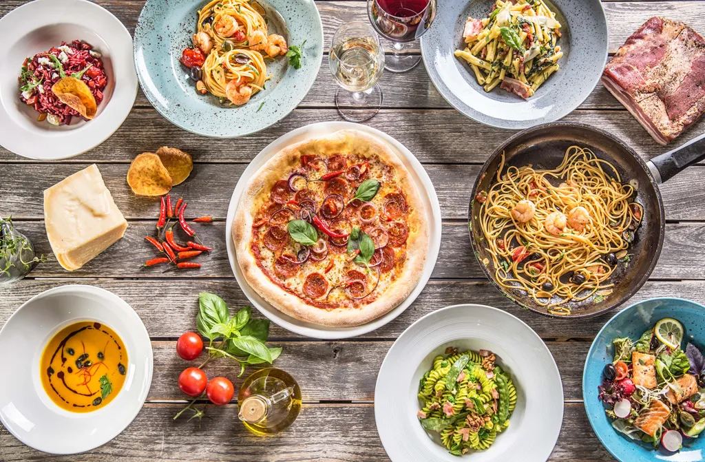 Verschiedene italienische Gerichte auf einem Holztisch, darunter Pizza, Pasta, Salate und Gläser Wein, umgeben von frischen Zutaten.