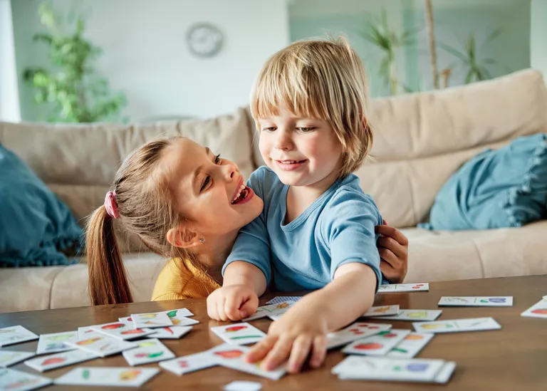 Zwei Kinder spielen fröhlich mit Bildkarten auf einem Tisch, umgeben von einer gemütlichen Wohnzimmerkulisse.