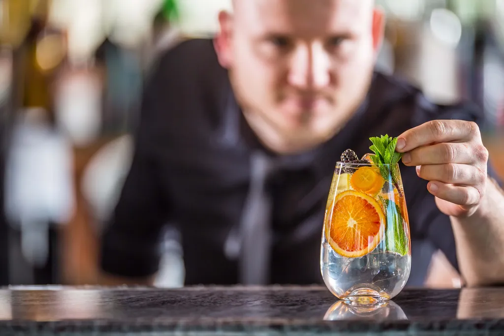 Eine Person garniert einen Cocktail mit Minze, Orangenscheiben und gelben Zitrusfrüchten in einem klaren Glas auf einer Bartheke.