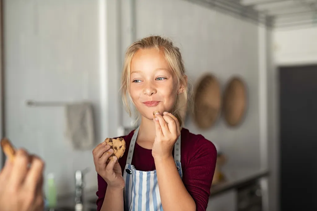 Ein Mädchen mit gestreifter Schürze nascht einen selbstgebackenen Keks in der Küche und hält einen weiteren Keks in der Hand.