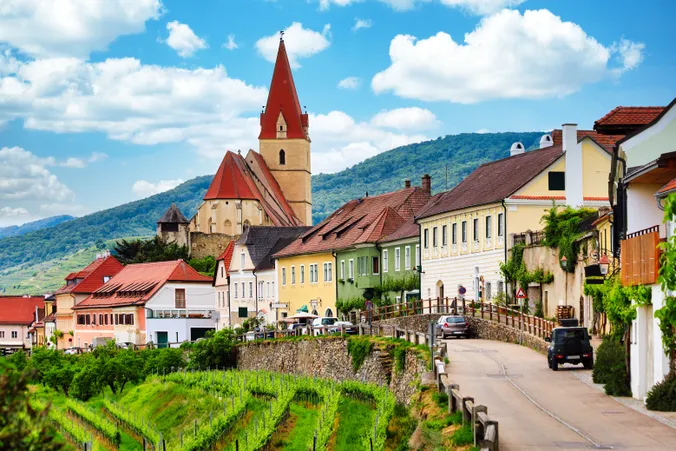 A picturesque village with colorful houses and a church with a red roof, set against a backdrop of green hills and a clear blue sky.