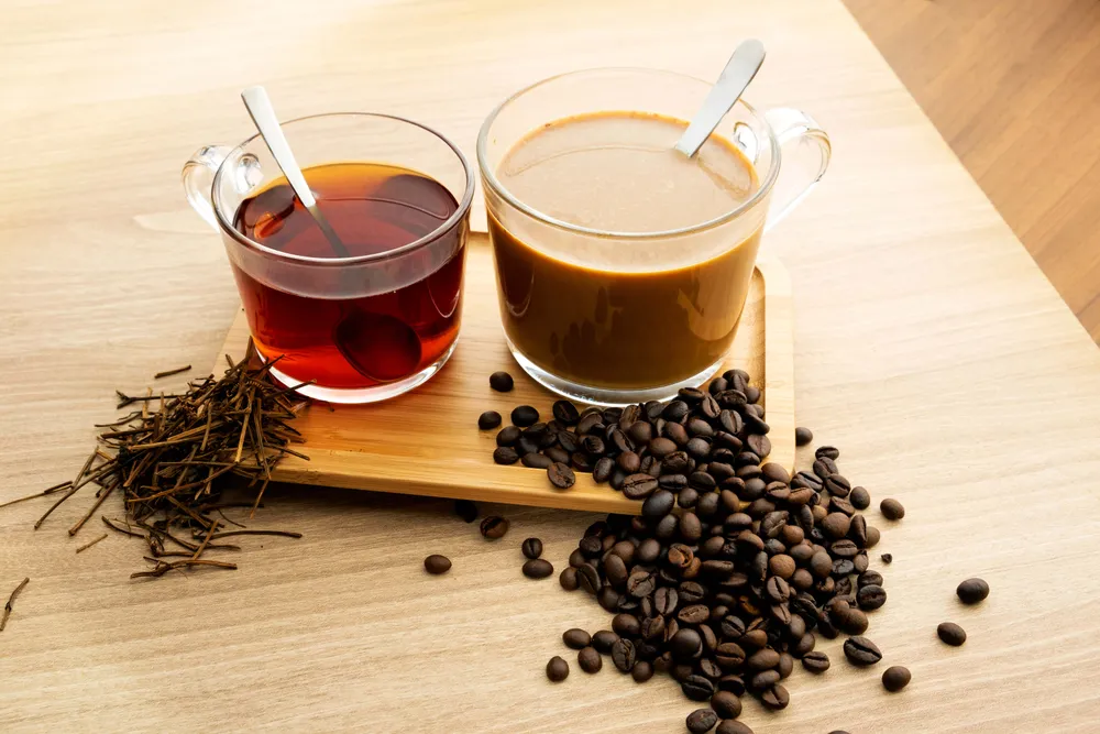 Zwei Glasbecher auf einem Holztisch; einer mit dunklem Tee, einer mit hellem Kaffee. In jedem Becher liegt ein silberner Löffel.