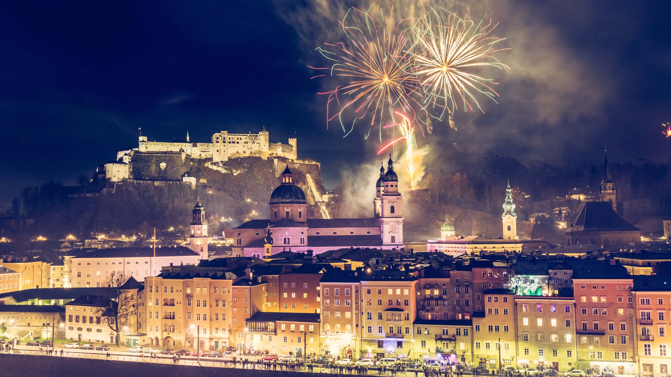 Ein Feuerwerk erhellt den Nachthimmel über einer historischen Stadtkulisse, im Hintergrund sind beleuchtete Gebäude und eine Festung auf einem Hügel zu sehen.
