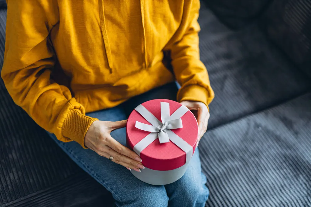 Person hält eine runde Geschenkbox mit rotem Deckel und weißer Schleife auf dem Schoß