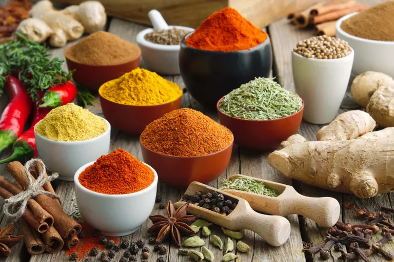 Eine Auswahl bunter Gewürze in Schälchen, darunter Kurkuma, Paprika und Rosmarin, die zusammen mit Ingwer, Chilischoten und Zimt auf einem Holztisch angeordnet sind.