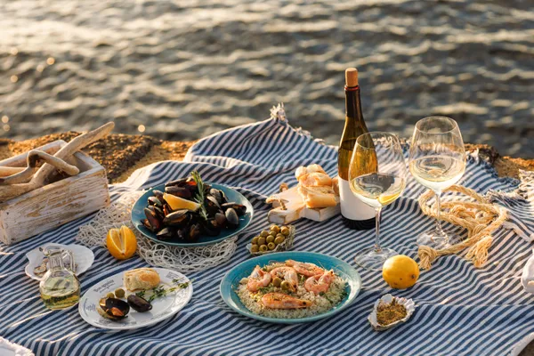 Picknick am Meer mit Meeresfrüchten, Brot, Oliven, Zitrone, einer Weinflasche und zwei Gläsern auf einer blau gestreiften Tischdecke am Wasser.