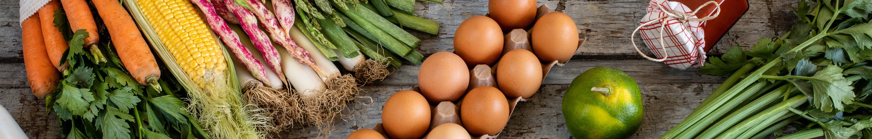 Auf einem rustikalen Holztisch liegen verschiedene frische Gemüsesorten, Obst, Eier und Brot. In einem Netzbeutel befinden sich einige Lebensmittel, außerdem stehen dort Flaschen.