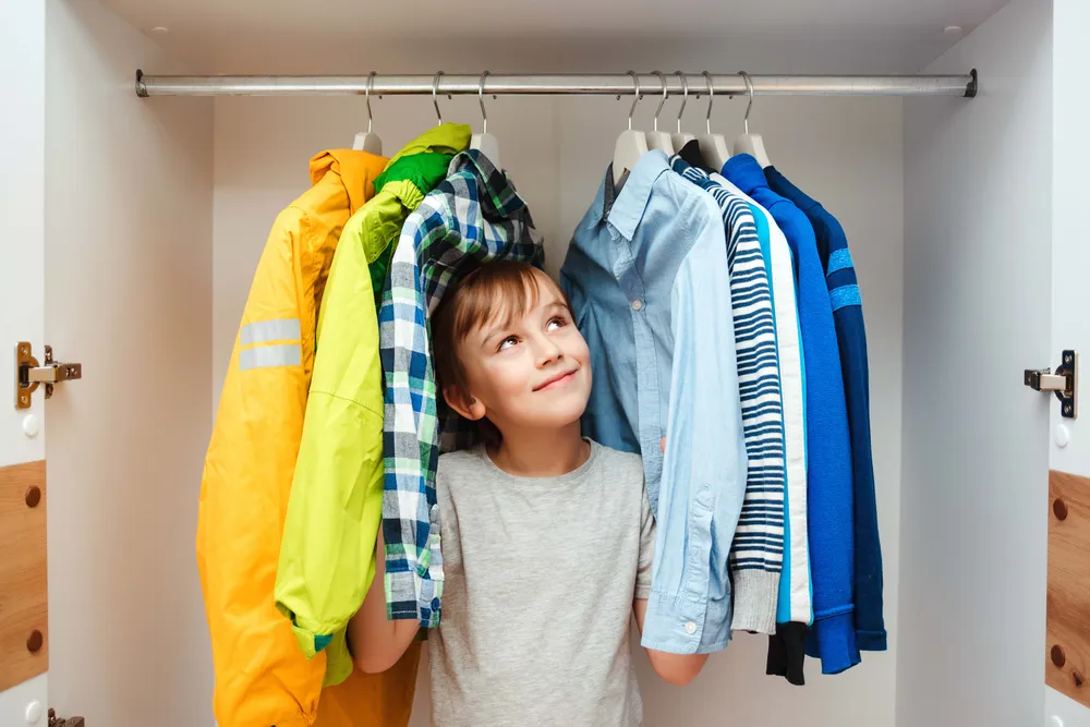 Kind steht zwischen aufgehängter Kinderkleidung in einem geöffneten Kleiderschrank