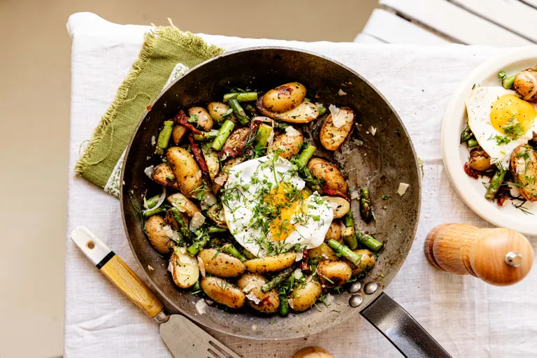 In einer Pfanne gebratene Kartoffeln, Spargel und Speck, garniert mit einem Spiegelei und Kräutern, auf einer weißen Tischdecke, daneben eine hölzerne Pfeffermühle.
