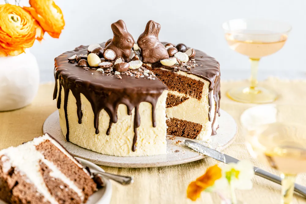 Mehrschichtiger Schokoladenkuchen mit Schokoladenhasen, Schokoladenglasur und Blumen, daneben zwei Gläser Champagner.