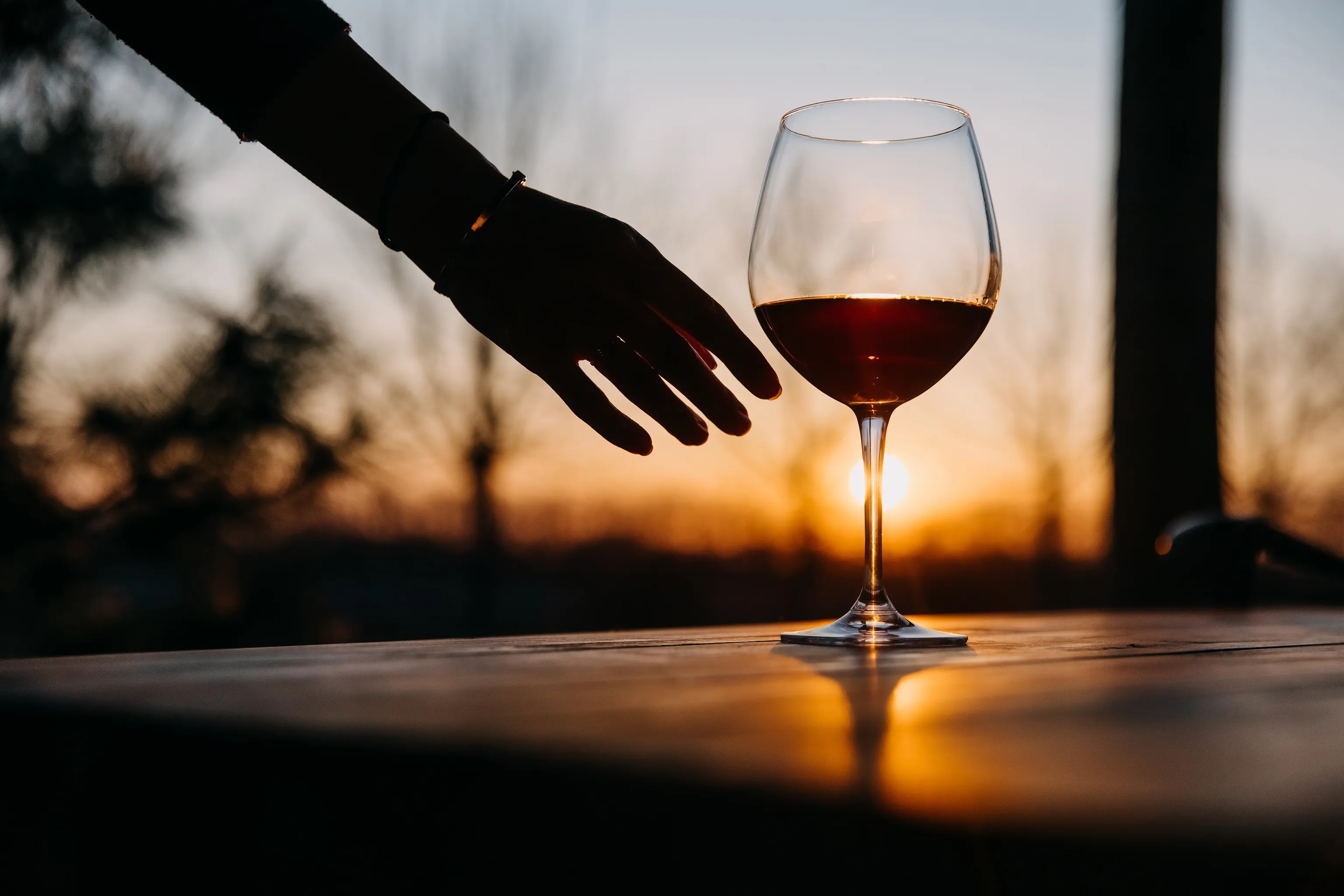 Eine Hand, die nach einem Weinglas auf einem Tisch greift, vor dem Hintergrund eines Sonnenuntergangs als Silhouette zu sehen.