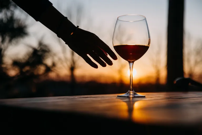 Eine Hand, die nach einem Weinglas auf einem Tisch greift, vor dem Hintergrund eines Sonnenuntergangs als Silhouette zu sehen.