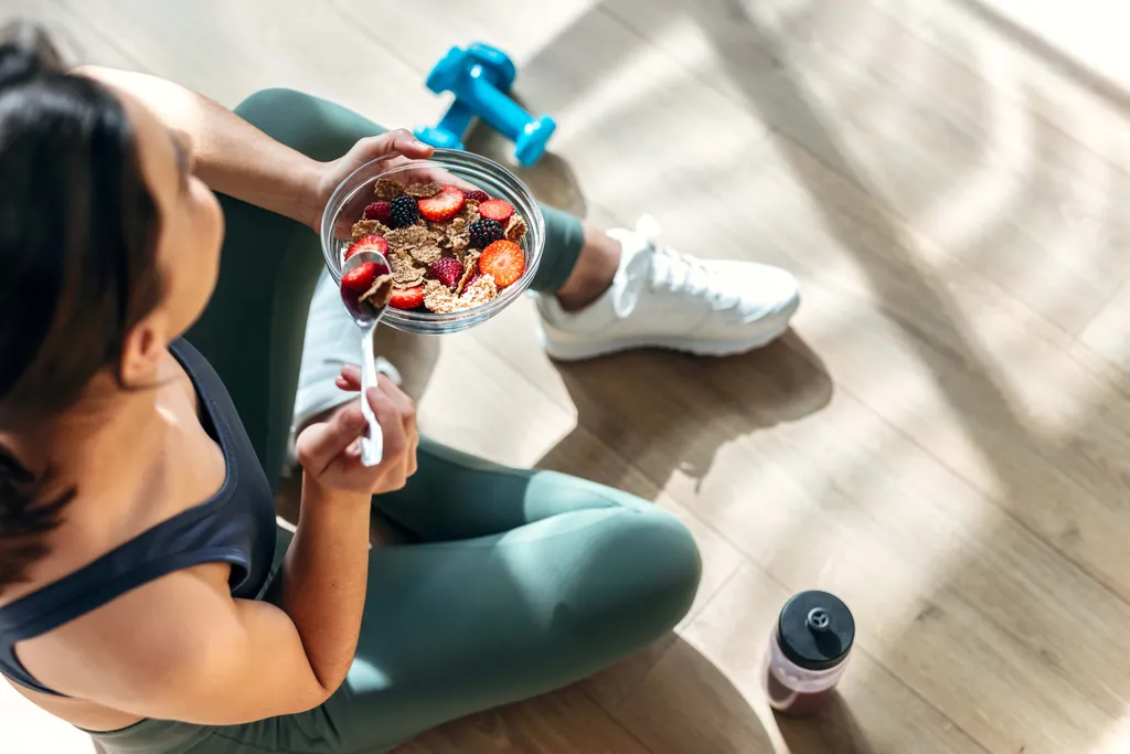 Eine Person in Sportkleidung sitzt auf dem Boden und isst eine Schüssel Müsli mit Beeren; daneben stehen eine Wasserflasche und eine Hantel.
