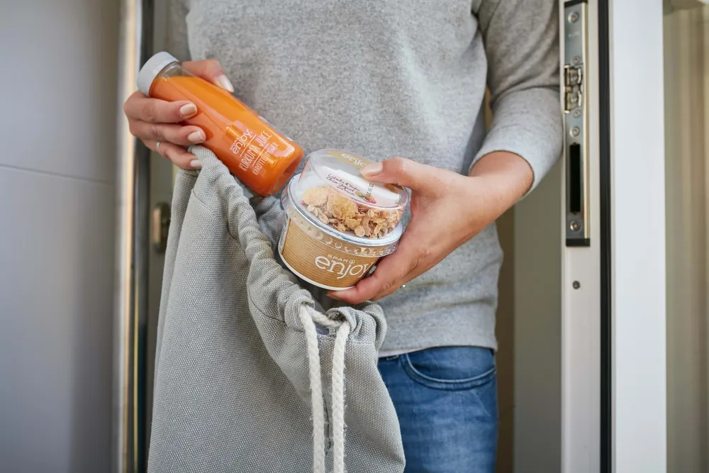 Eine Person in einem grauen Pullover hält eine Flasche Karottensaft und einen Becher Müsli in der Hand und steckt beides in einen grauen Beutel mit Kordelzug.