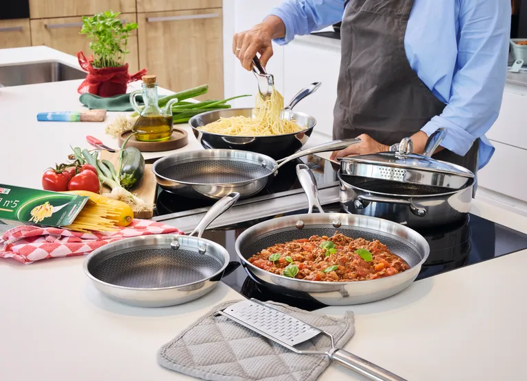 Unterschiedliche große Pfannen mit einer Wabenbeschichtung, die das Anhaften von Speisen verhindert. Diese stehen auf einer Kücheninsel und jemand kocht gerade Spaghetti Bolognese.