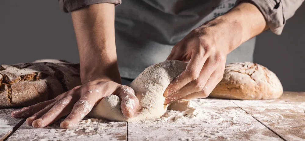 Hände, die auf einer bemehlten Arbeitsfläche Teig kneten, im Hintergrund frisch gebackenes Brot.