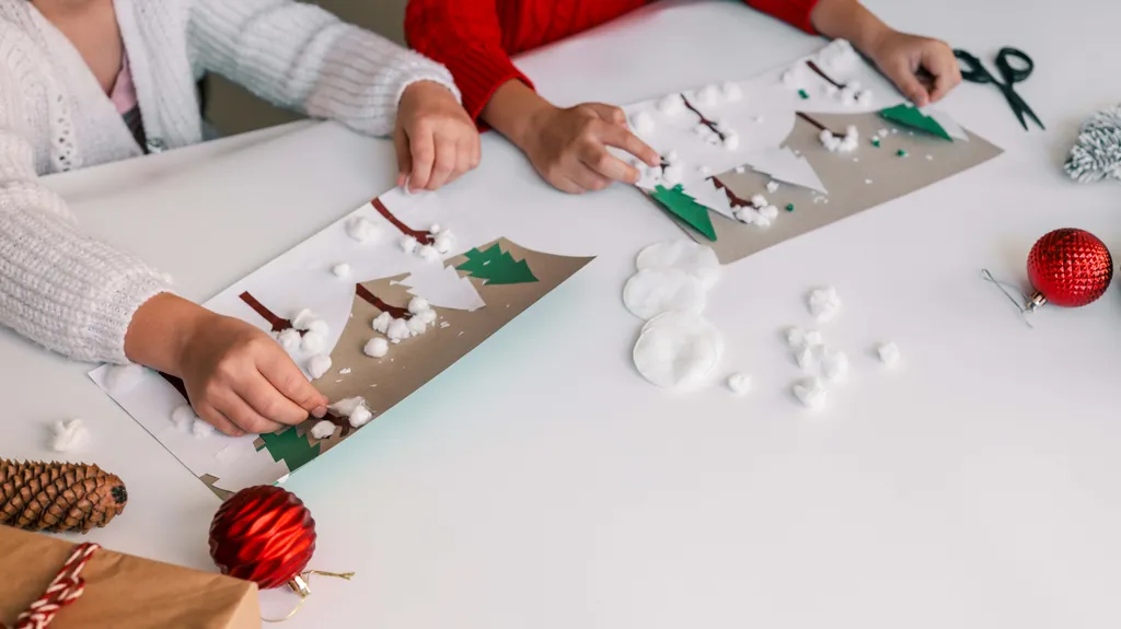 Kinder basteln Winterszenen aus Papier und Wattebällchen, umgeben von festlicher Dekoration wie Tannenzapfen und einem roten Christbaumschmuck.