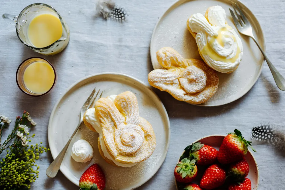 Zwei mit Puderzucker bestäubte Windbeutel auf Tellern, umgeben von Erdbeeren, einem Glas Vanillecreme und Blumen auf einer hellen Tischdecke.