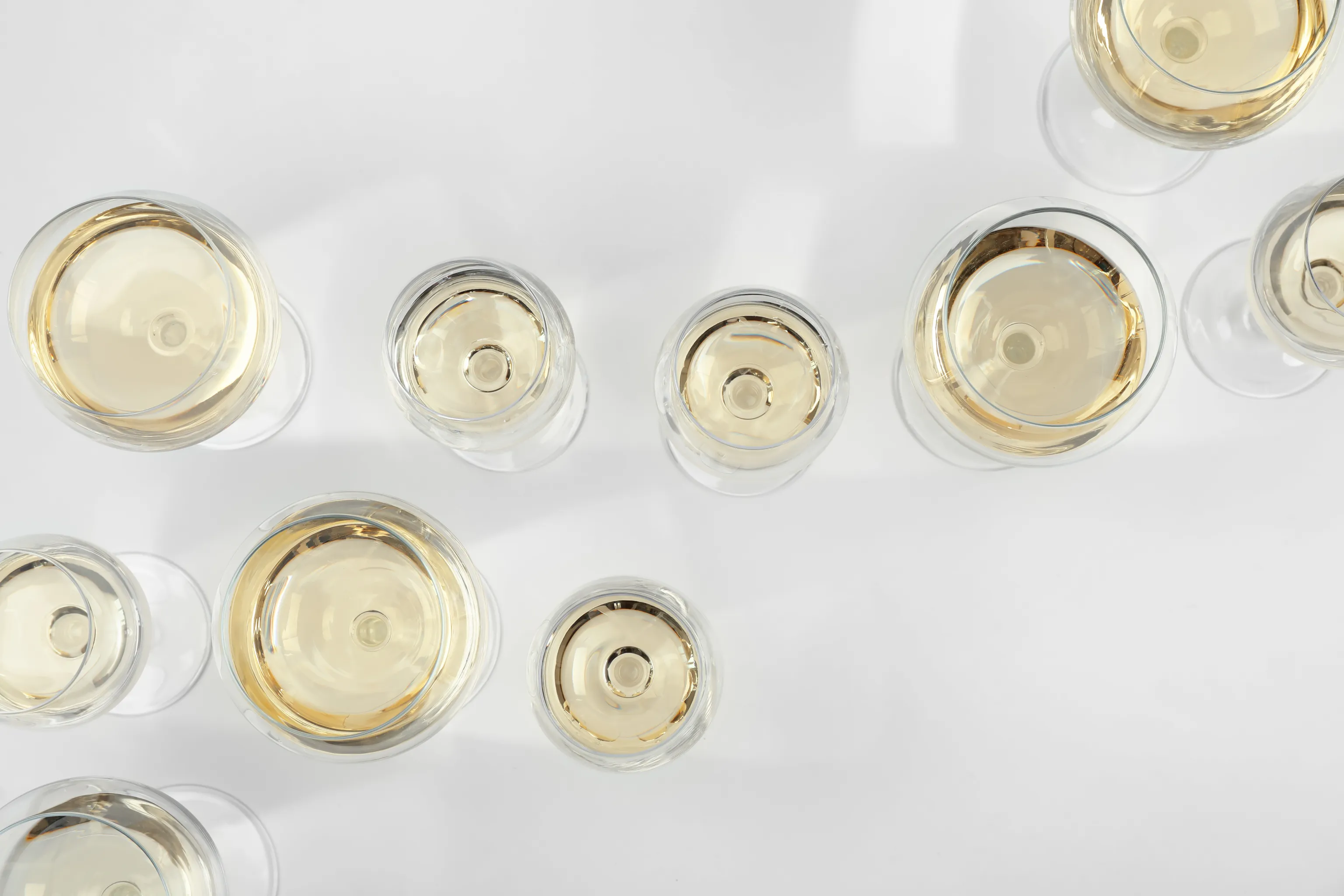 Neun Gläser Weißwein, von oben betrachtet auf einer weißen Fläche, die sanfte Schatten werfen.