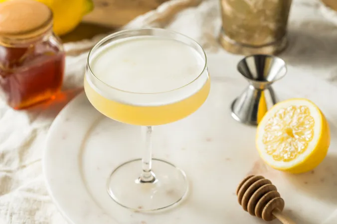 Ein Coupé-Glas, gefüllt mit einem schaumigen, gelben Cocktail, umgeben von Honig, einer Zitronenhälfte, einem Jigger und einem Honiglöffel auf einem weißen Tablett.