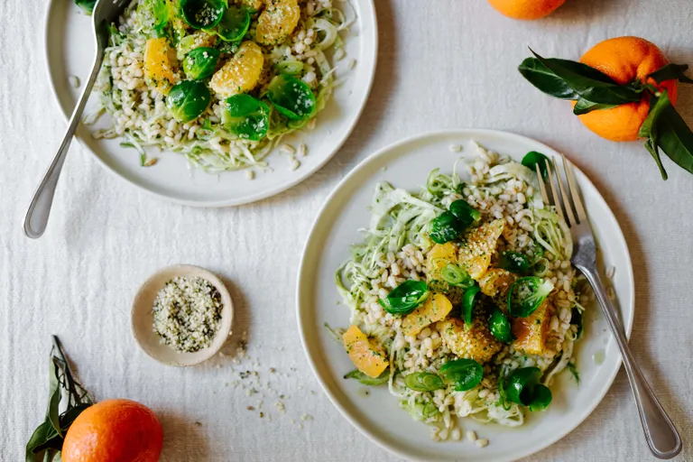 Zwei Teller mit frischem Salat aus Zucchininudeln, Orangenscheiben, Rosenkohl und Samen, serviert mit Orangen und einer kleinen Schale mit Samen.